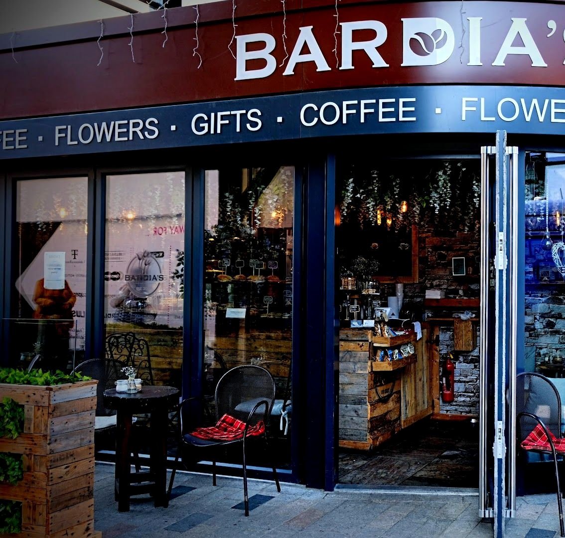 A Bardia's shopfront with a sign reading "Flowers · Gifts · Coffee", plants in a wooden planter and café chairs outside